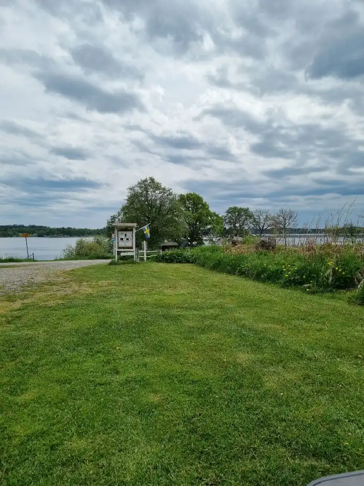 En grön gräsmatta breder ut sig under en blå himmel med moln, omgiven av frodig växtlighet. I bakgrunden syns en kustlinje med utsikt över ett öppet hav, som bidrar till platsens natursköna omgivning vid Säms camping och café.