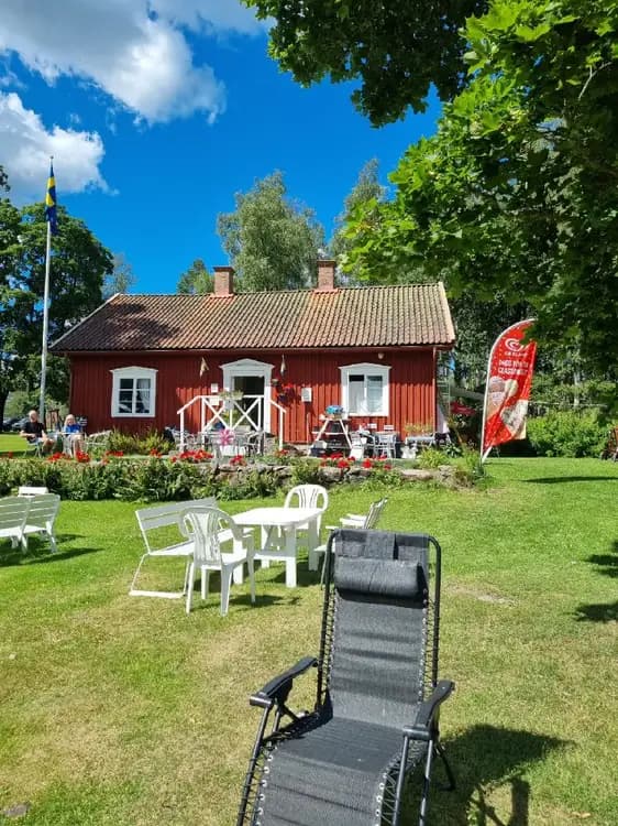 En mysig campingplats med grönskande växter och en välskött gräsmatta. Utemöbler som inkluderar bord och stolar står på gården vid en charmig stuga, vilket skapar en inbjudande miljö i skuggan.