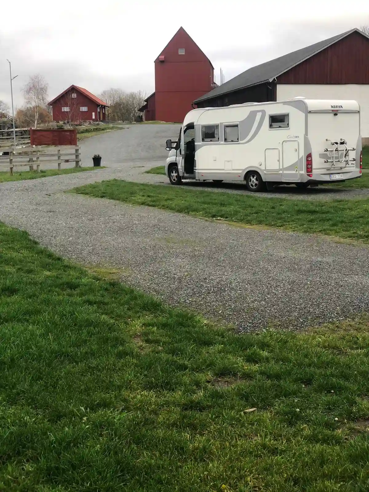En grusväg leder fram till en grön gräsmatta där flera husbilar och husvagnar är parkerade. En liten bil står närmast kameran. Platsen är Kedumsvik ställplats, omgiven av natur.