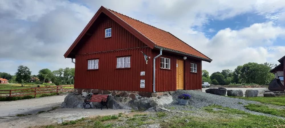 Bild av en träbyggnad på Kedumsvik ställplats. Byggnaden liknar en stuga med trätak och väggar, belägen i lantlig miljö. En dörr syns på framsidan av byggnaden.