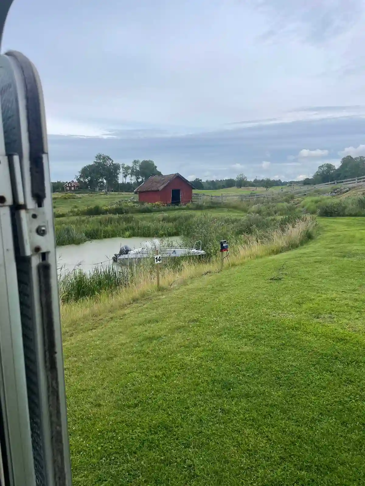En vidsträckt och grönskande landskap med blå himmel och vita moln över Kedumsvik ställplats. Det öppna fältet omges av frodigt gräs och växter, vilket skapar en fridfull och lantlig atmosfär.