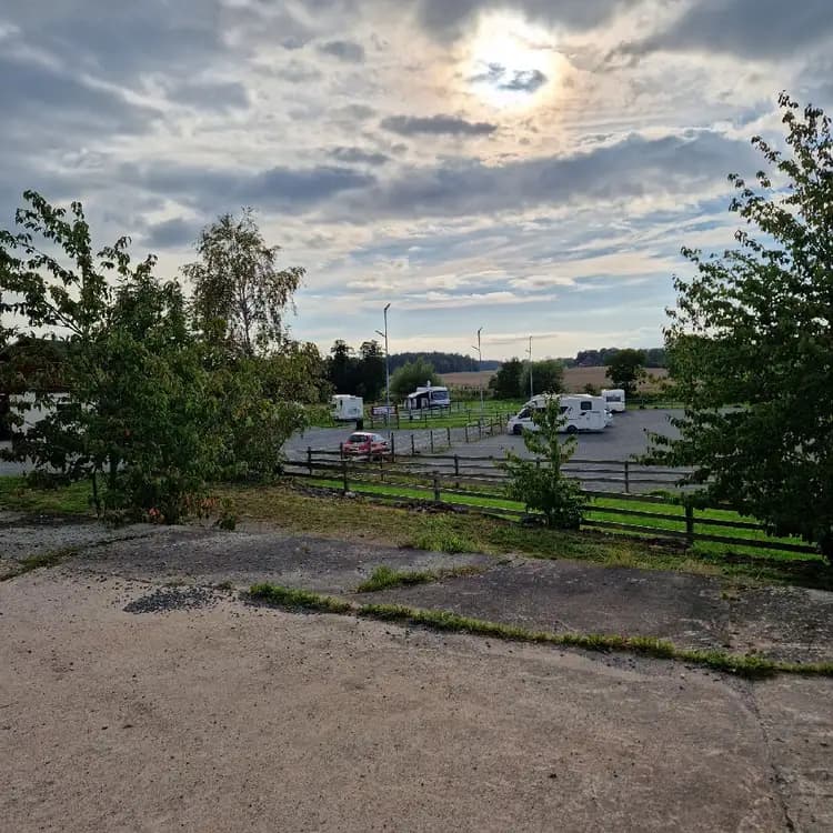 En familjebil och en lyxbil parkerade på Kedumsvik ställplats under en klar dag med några cumulusmoln på himlen och gröna växter i bakgrunden.