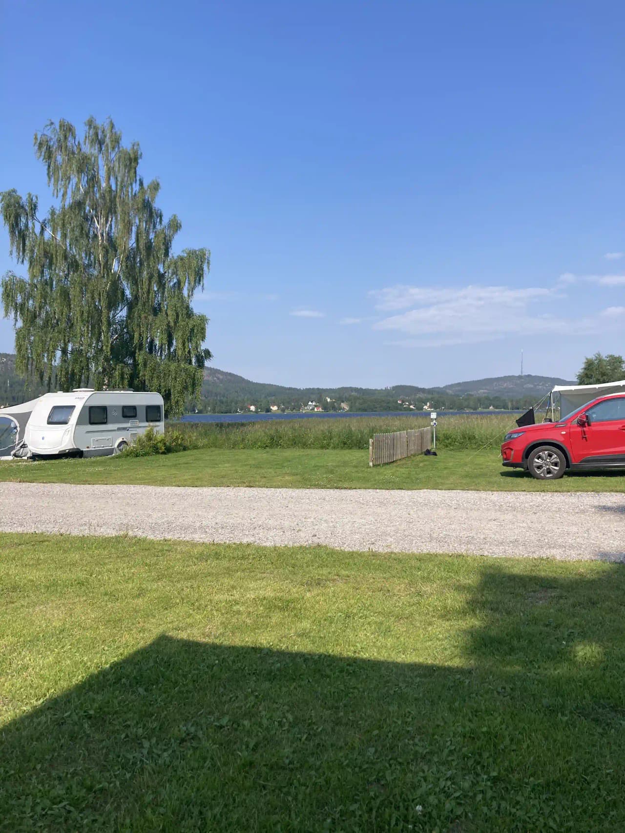 En grön campingplats där flera husbilar och fordon är parkerade på en gräsmatta. Det finns en blandning av familjebilar och lyxigare fordon på området.