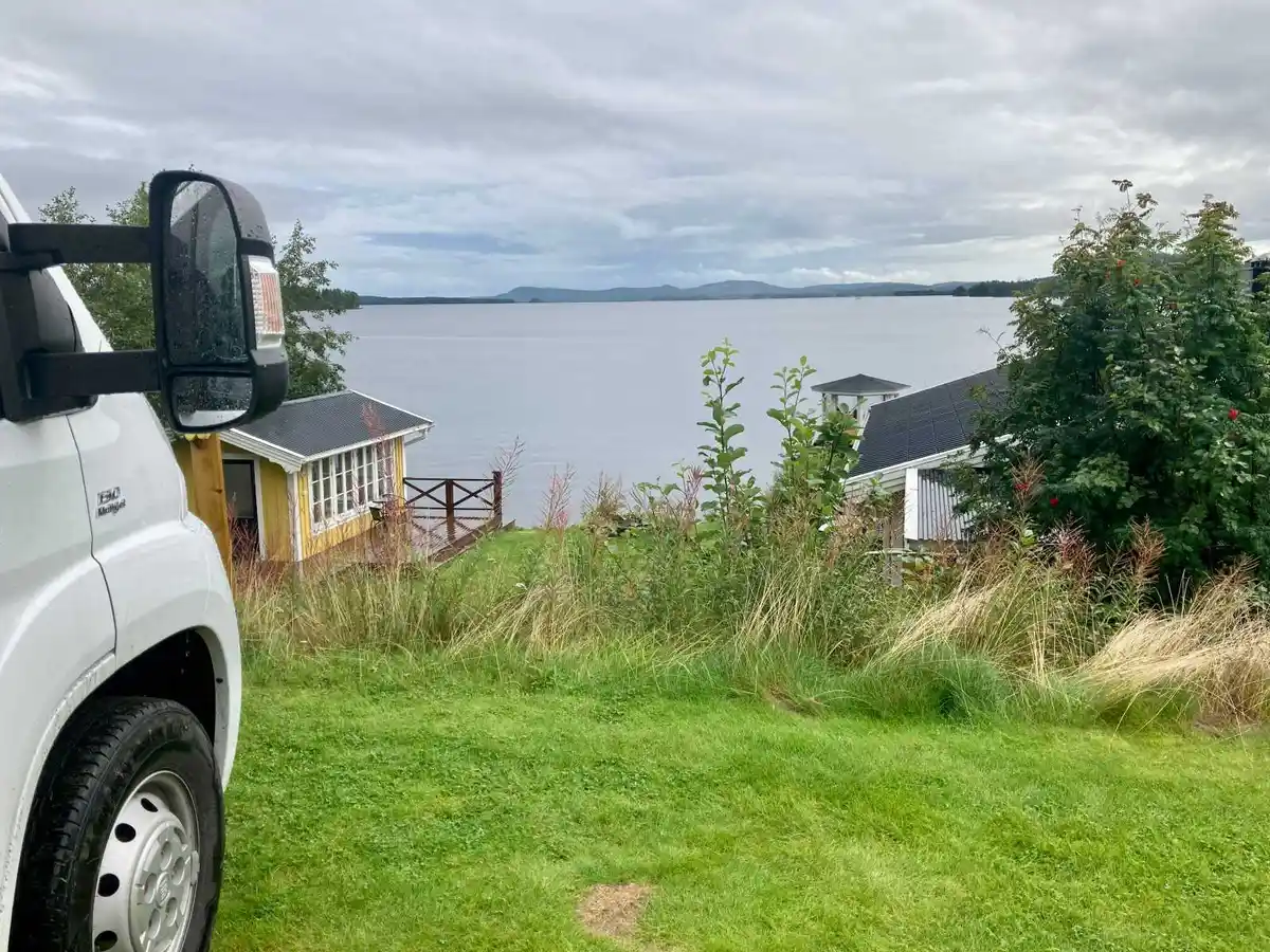 En grönskande campingplats med en stuga och flera träd i bakgrunden. Himlen är delvis täckt av vita moln. En ljus kommersiell fordon står parkerad på gården.