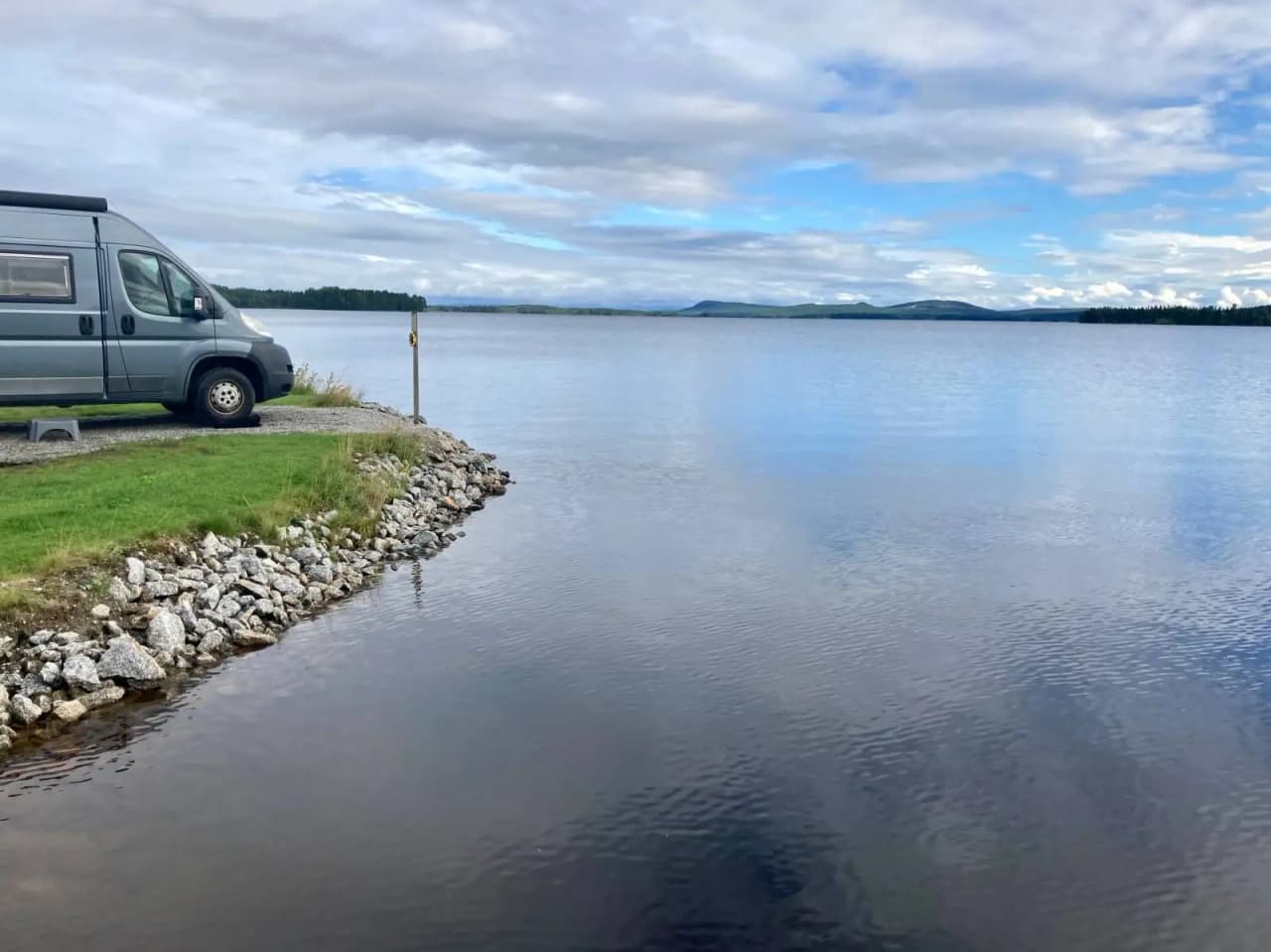 En lugn sjö med blått vatten under en lätt molnig himmel, omgiven av grönskande strandkanter vid Kolgårdens stugby & camping.