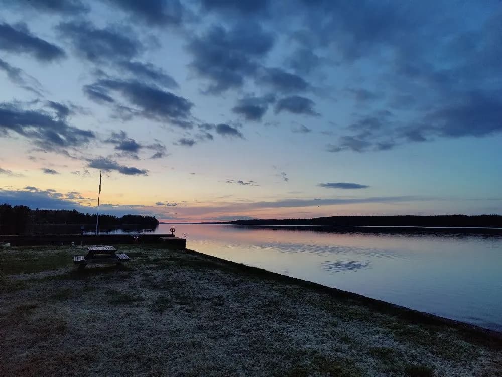 En campingplats vid Västanviksbadet i Leksand med utsikt över en vattenyta. Himlen är blå med lätta moln vid horisonten och solen går ner vid skymning.