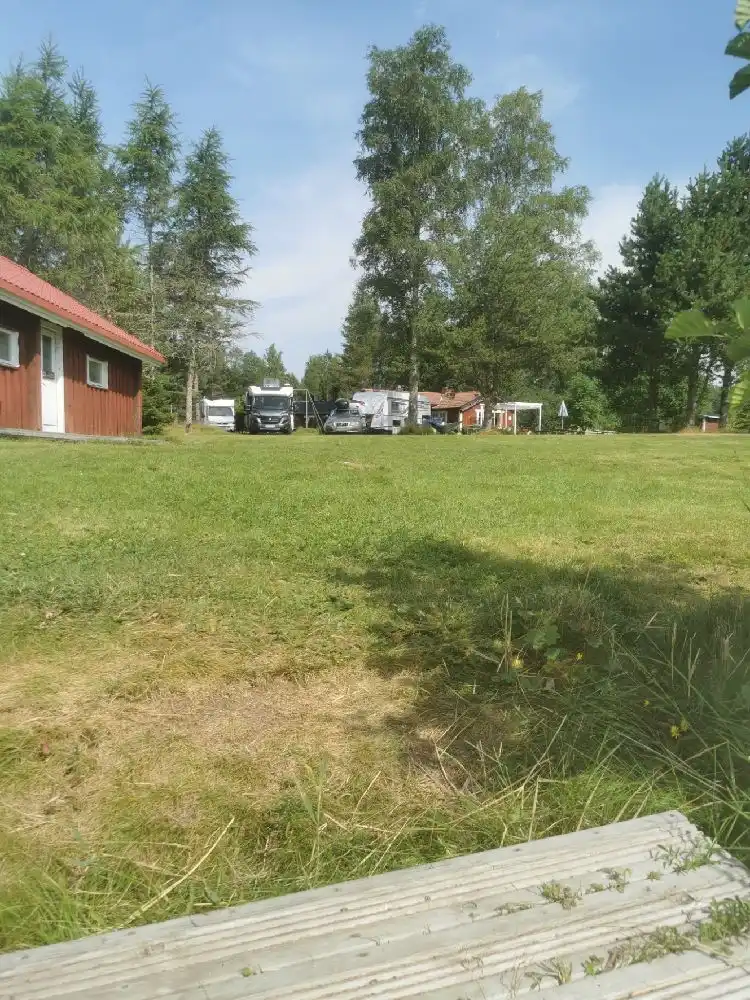 En grönskande campingplats med gräsmattor, omgiven av träd och växtlighet. Små stugor ligger utspridda i ett naturskönt, lantligt område.