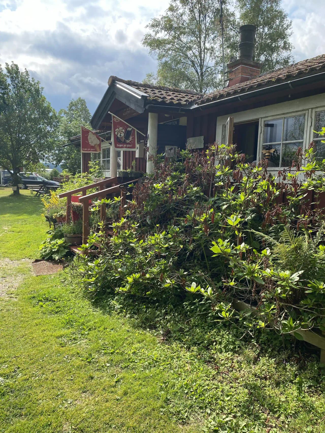 En vit stuga omgiven av grönskande buskar och gräsmatta på Lesjöbyns camping, med ett ordnat trädgårdslandskap i förgrunden.
