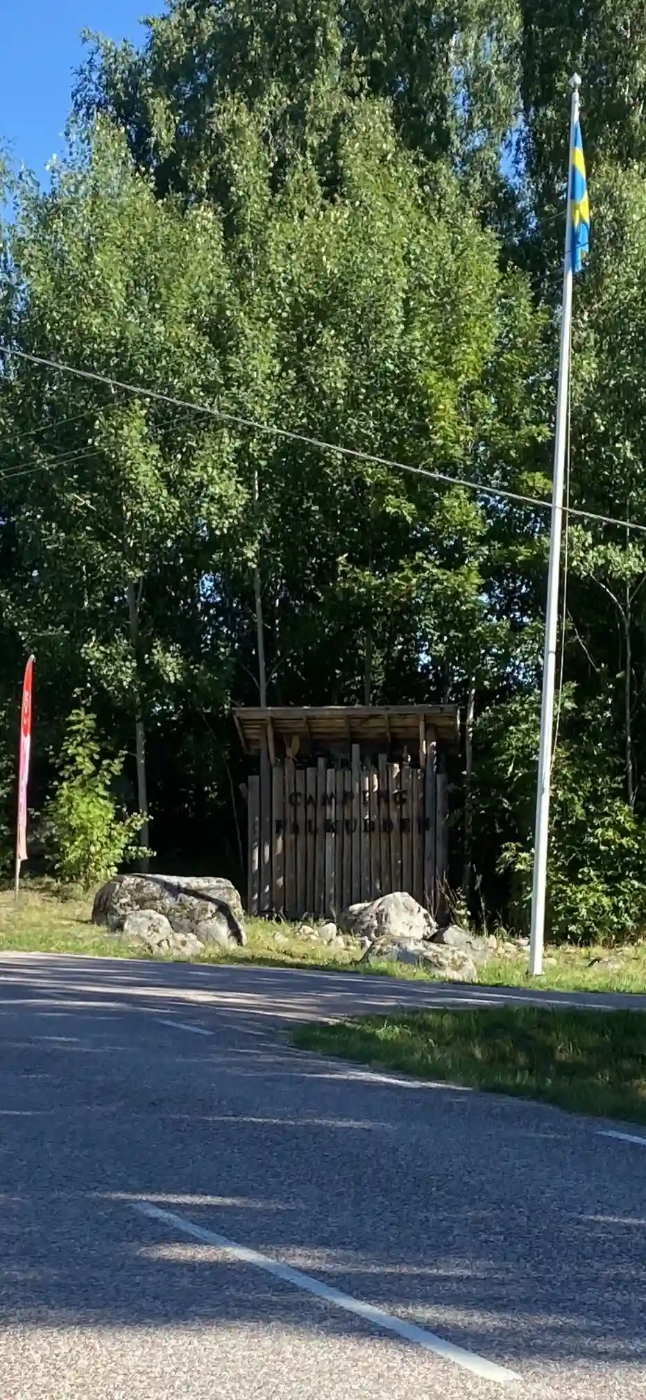 En skogsområde med höga träd, en grusväg leder in i området. Det finns en flaggstång med svenska flaggan vid sidan av vägen. Skugga från trädkronorna täcker delar av stigen.