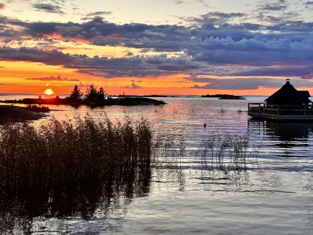En lugn sjövid Filsbäcks camping i skymningen, med spegelblanka vattenytor som reflekterar den färgstarka solnedgången vid horisonten.