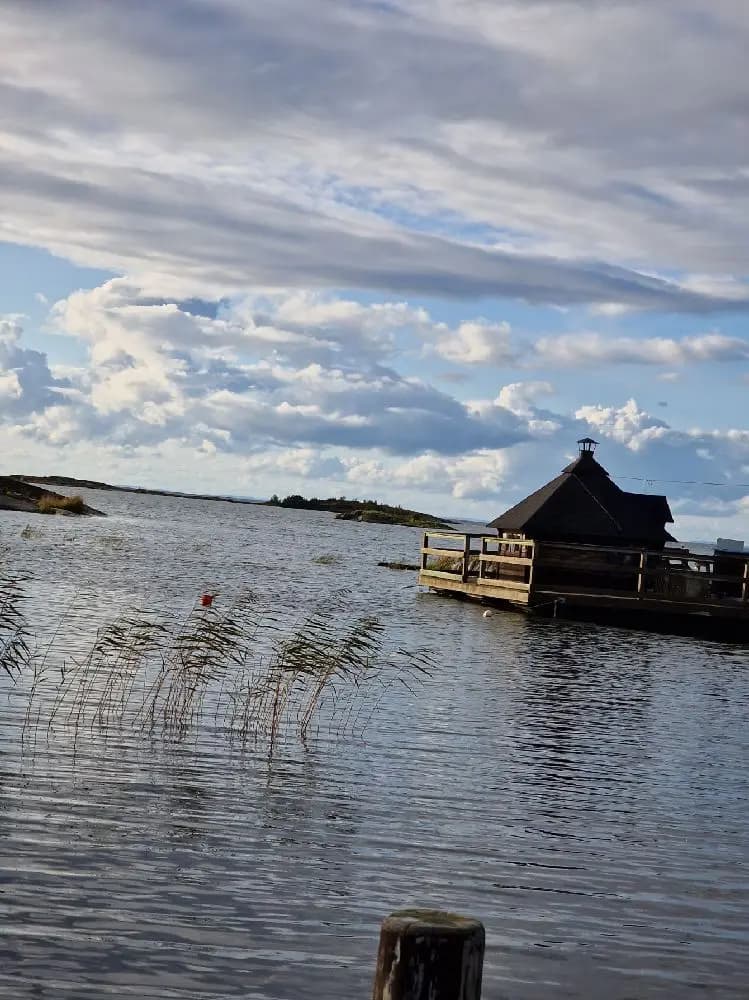 Bild av Filsbäcks camping vid en sjö med molnig himmel. En liten stuga med halmtak syns nära vattnet, omgiven av våtmarker och grönska.