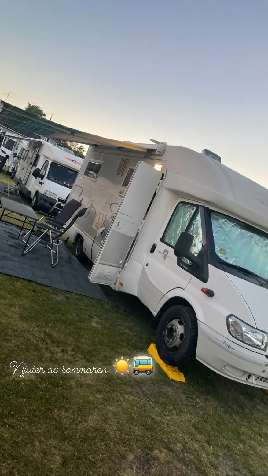 En husbil står parkerad på Åsa Camping & Havsbad, omgiven av grönska och träd. Temperaturen är behaglig och det är solsken. Det syns också några andra fordon i bakgrunden.