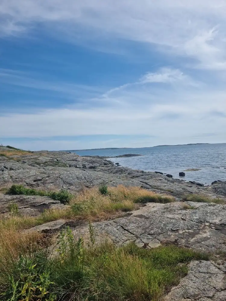 En vy över Åsa Camping & Havsbad med blå himmel, horisont och kustlinje. Stenar och klippor vid havet, som sträcker sig ut i det öppna havet under en spridd molnig himmel.