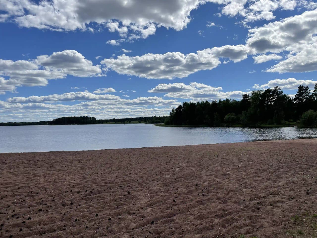 En vy över Våmåbadets camping med en blå himmel och moln i bakgrunden. Vattnet sträcker sig över bildens mitt, med strandlinjen synlig i fjärran.