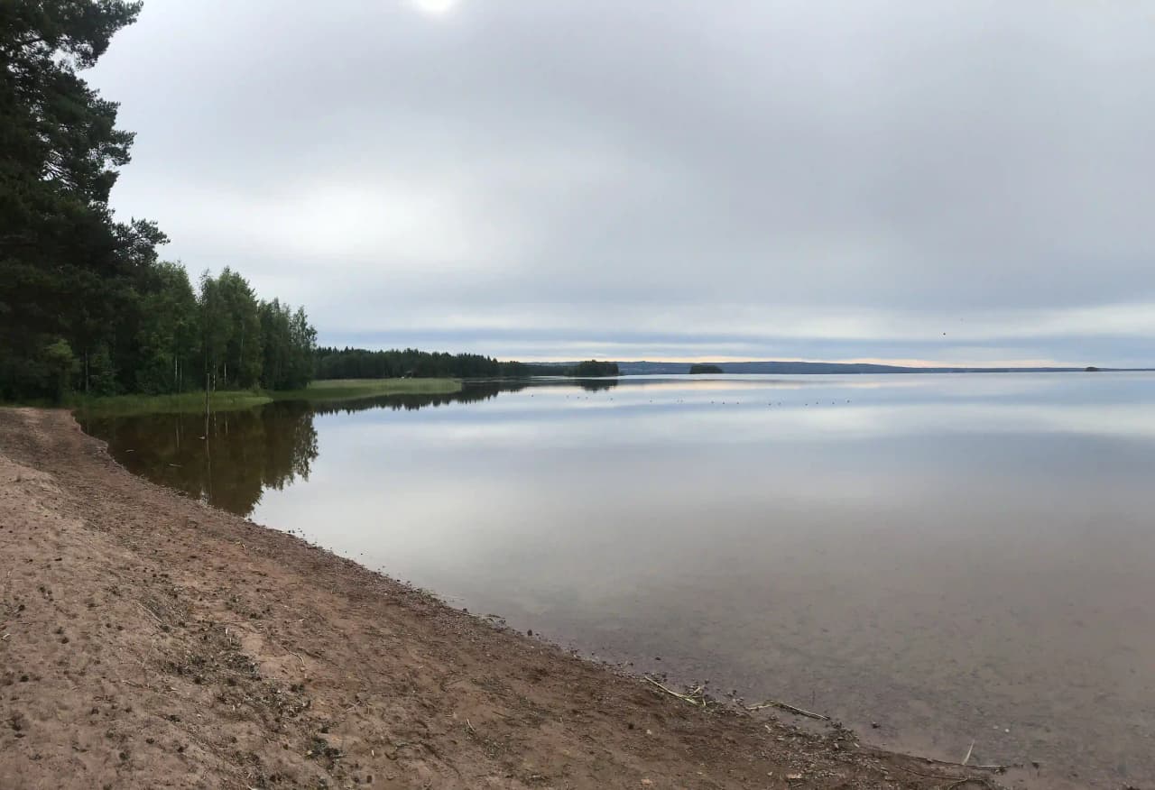 En vacker bild av Våmåbadets camping med ett vidsträckt vattendrag i förgrunden. Moln skimrar på horisonten och skapar en fridfull atmosfär vid den natursköna stranden.