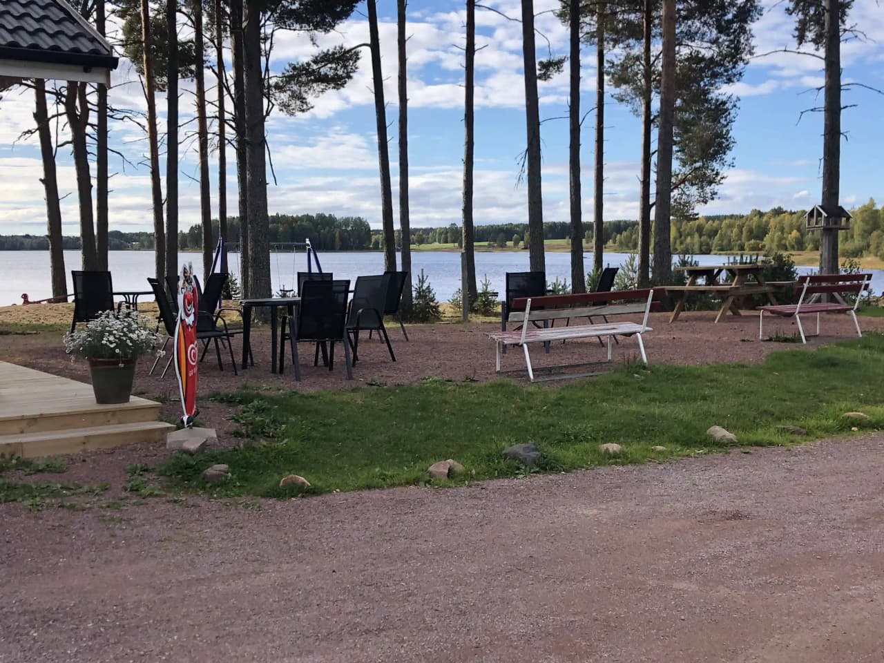 En campingplats vid strandkanten med skuggiga områden, utemöbler och en stolpe. I bakgrunden syns en sjö och omgivande naturlandskap.