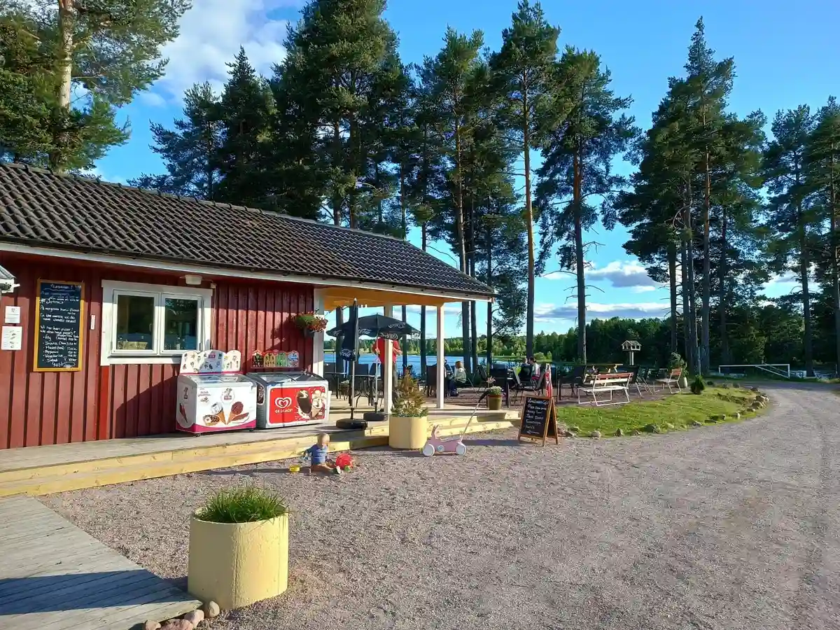 En campingplats vid Våmåbadet med stugor i bakgrunden, ett picknickbord under skugga och en sandig uppfart.