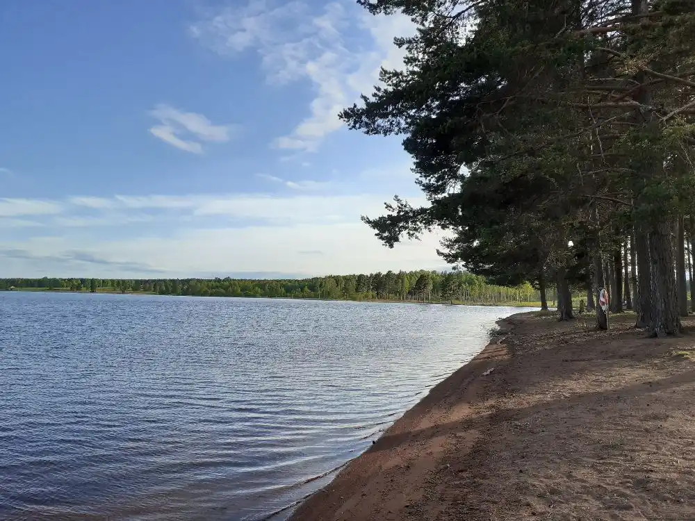 Bild av Våmåbadets camping med en klar blå himmel och moln i bakgrunden. Insjön sträcker sig mot horisonten, omgiven av grönskande natur och speglar den lugna, blå himlen.