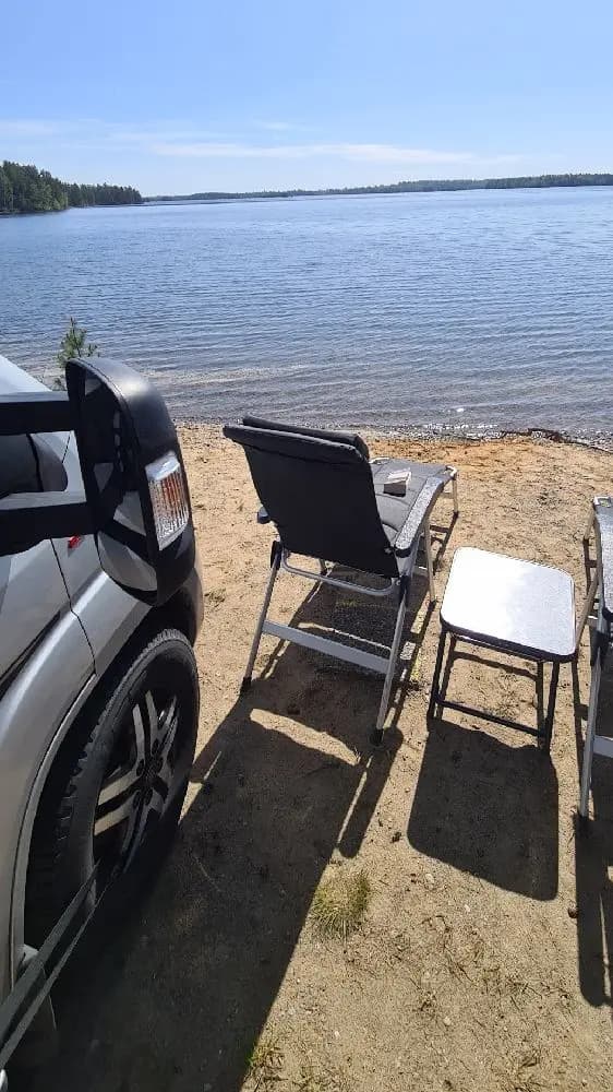 En öde strand på Revelbadets camping med fällstolar och ett bord vid sandkanten, utsikt mot bukten i bakgrunden.