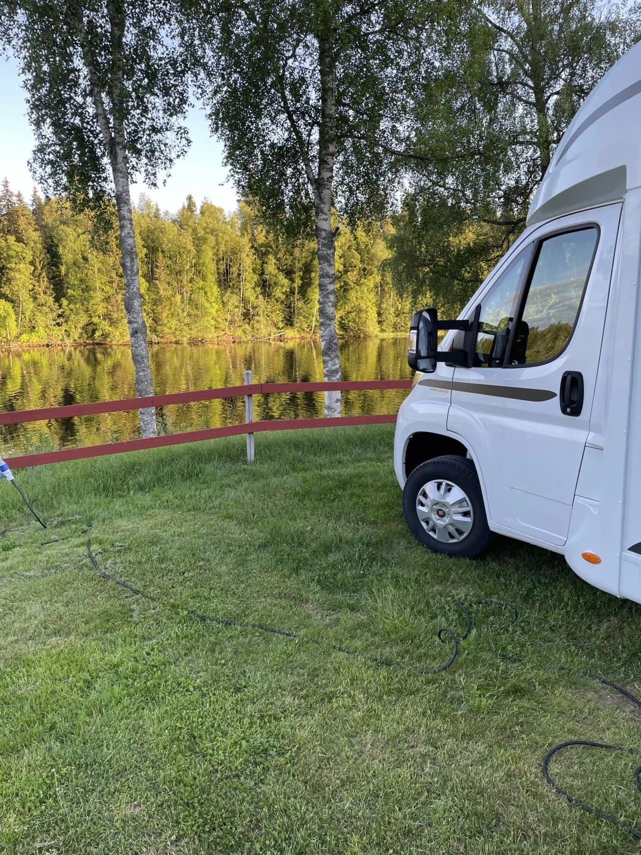 En grön gräsmatta med en parkerad vit minibuss vid sidan av en bil. Fjorden och skog kan anas i bakgrunden under en klar himmel.