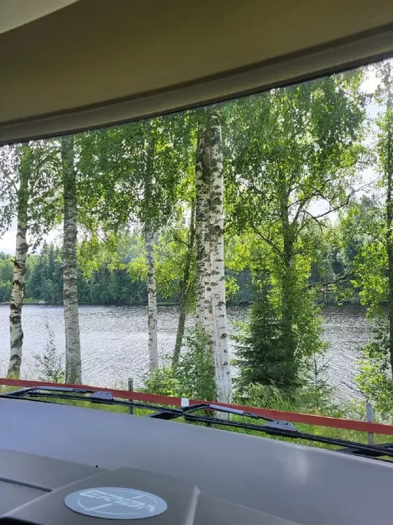 Bild av Grebbestadfjorden: En lugn fjord omgiven av tät skog med barrträd såsom lärk och gran. Solens strålar skapar vackra reflektioner på fjordens glasklara vatten, och skuggor spelar över landskapet. Det är en dag med klart ljus och man kan ana den rofyllda atmosfären i området.