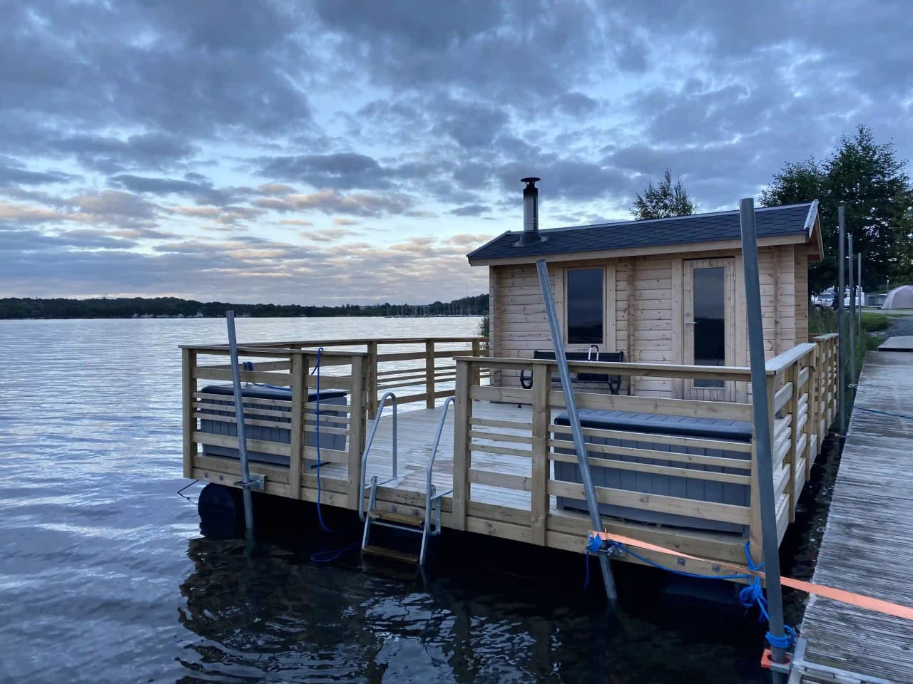 En träbrygga sträcker sig ut över en spegelblank sjö, omgiven av tät skog, vid Lövekulle camping.