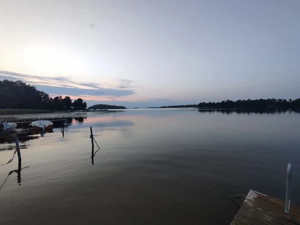 En stilla sjö med spegelblank vattenyta vid skymningen, omgiven av ett öppet landskap. Solnedgången sätter en varm ton över himlen och vattnet, vilket skapar en lugn och avkopplande atmosfär vid Sundbyholms camping.