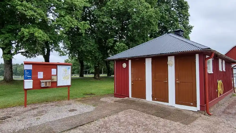 Alt-text: Hofsnäs Herrgård med trädgårdsbyggnader och en uppfart i skugga, omgiven av grönska i en pittoresk miljö.