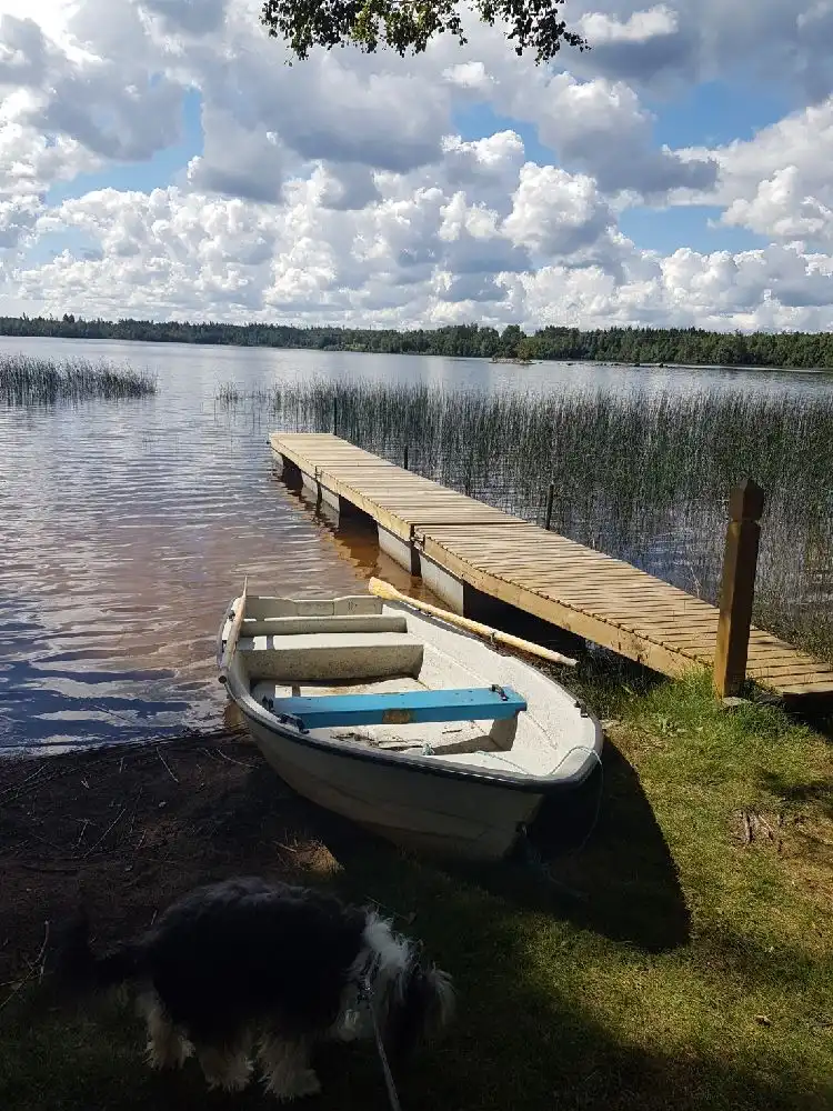 En idyllisk vy över ett spegelblankt vattendrag med små båtar förtöjda vid kanten, omgiven av grönskande natur. Moln pryder den blå himlen ovanför.