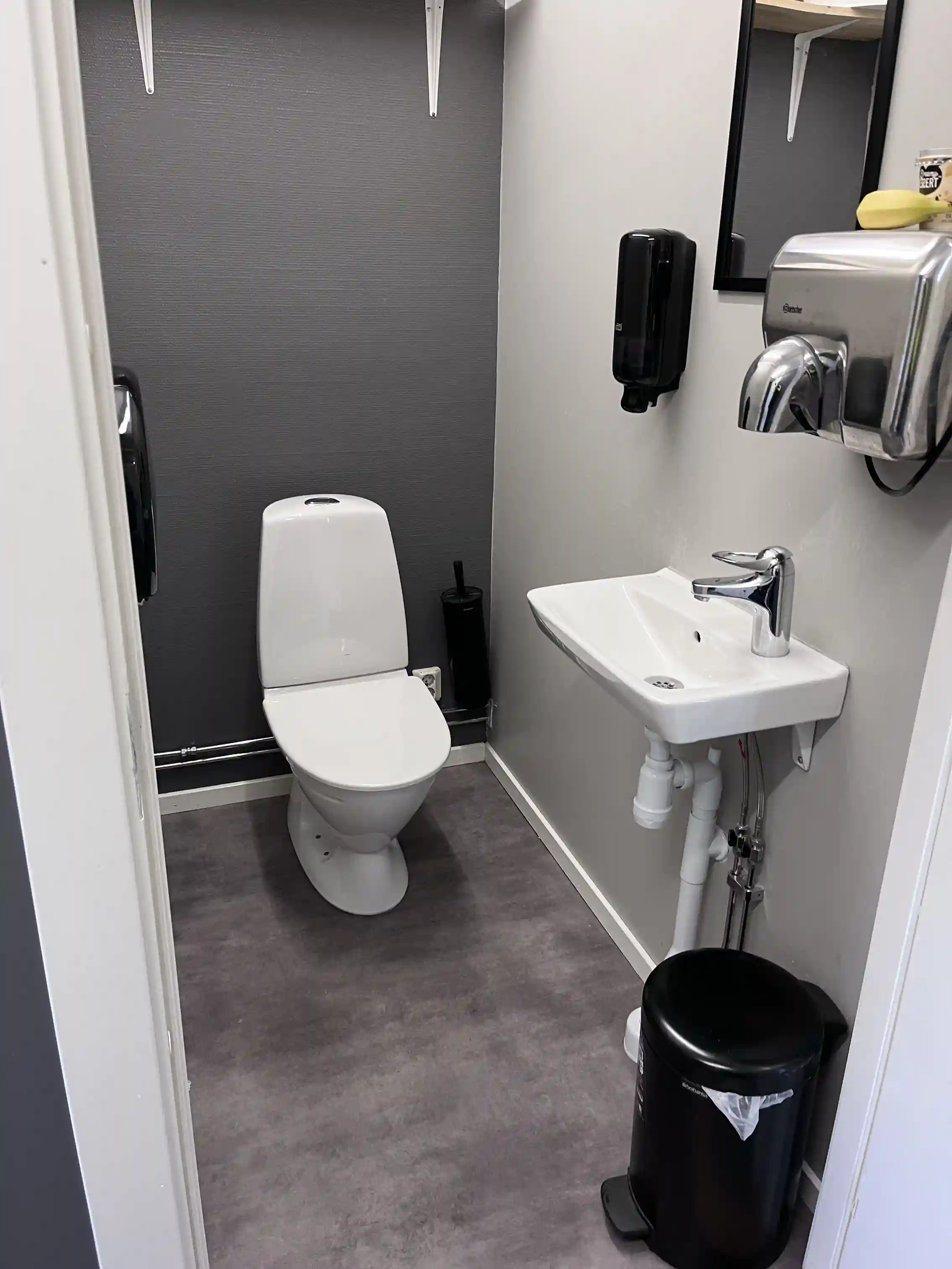 En bild av ett grått kaklat badrum med toalett, golvplattor och silverfärgade rör- och sanitetsarmaturer, på Arvika småbåtshamn.