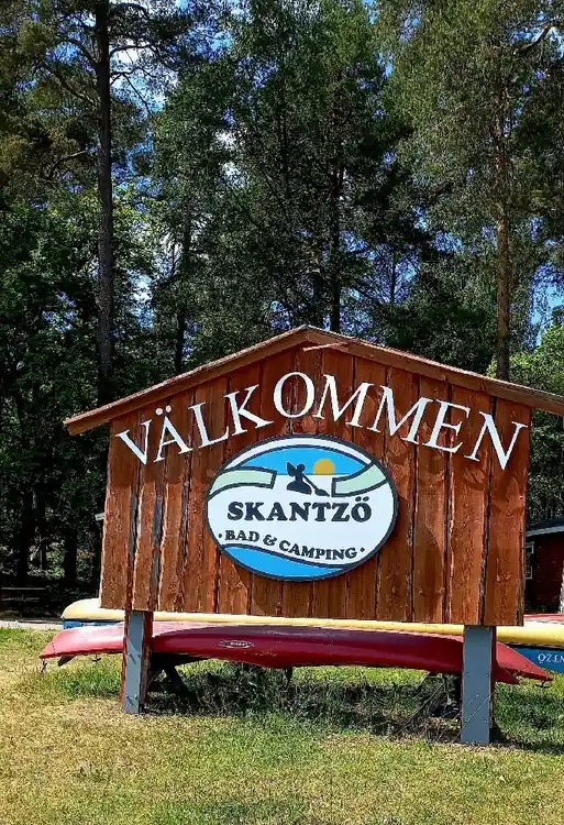 En träskylt vid ingången till Skantzö Bad & Camping, omgiven av grönska och träd, visar vägen till rekreationsområdet.