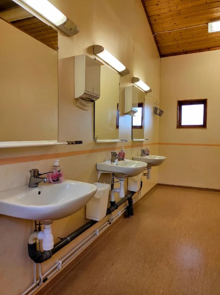 Badrum med ljusa väggar och ett klinkergolv. Ett handfat med kran och spegel ovanför, belyst av takbelysning. Rummet är enkelt och funktionellt designat.