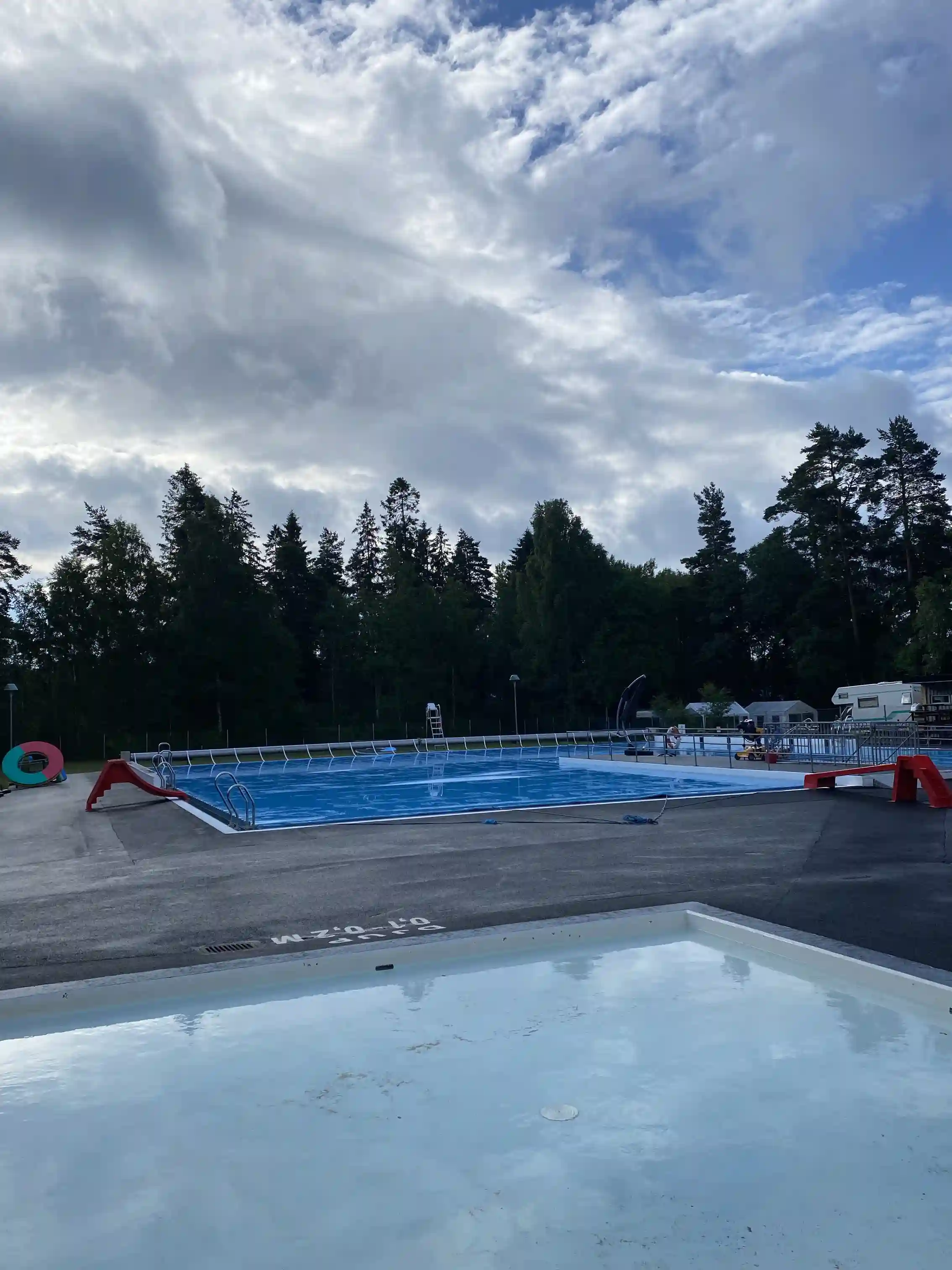 En bild av Hörsjöns camping med tallar i bakgrunden, en pool för bad och avkoppling, under en himmel med vita cumulusmoln.