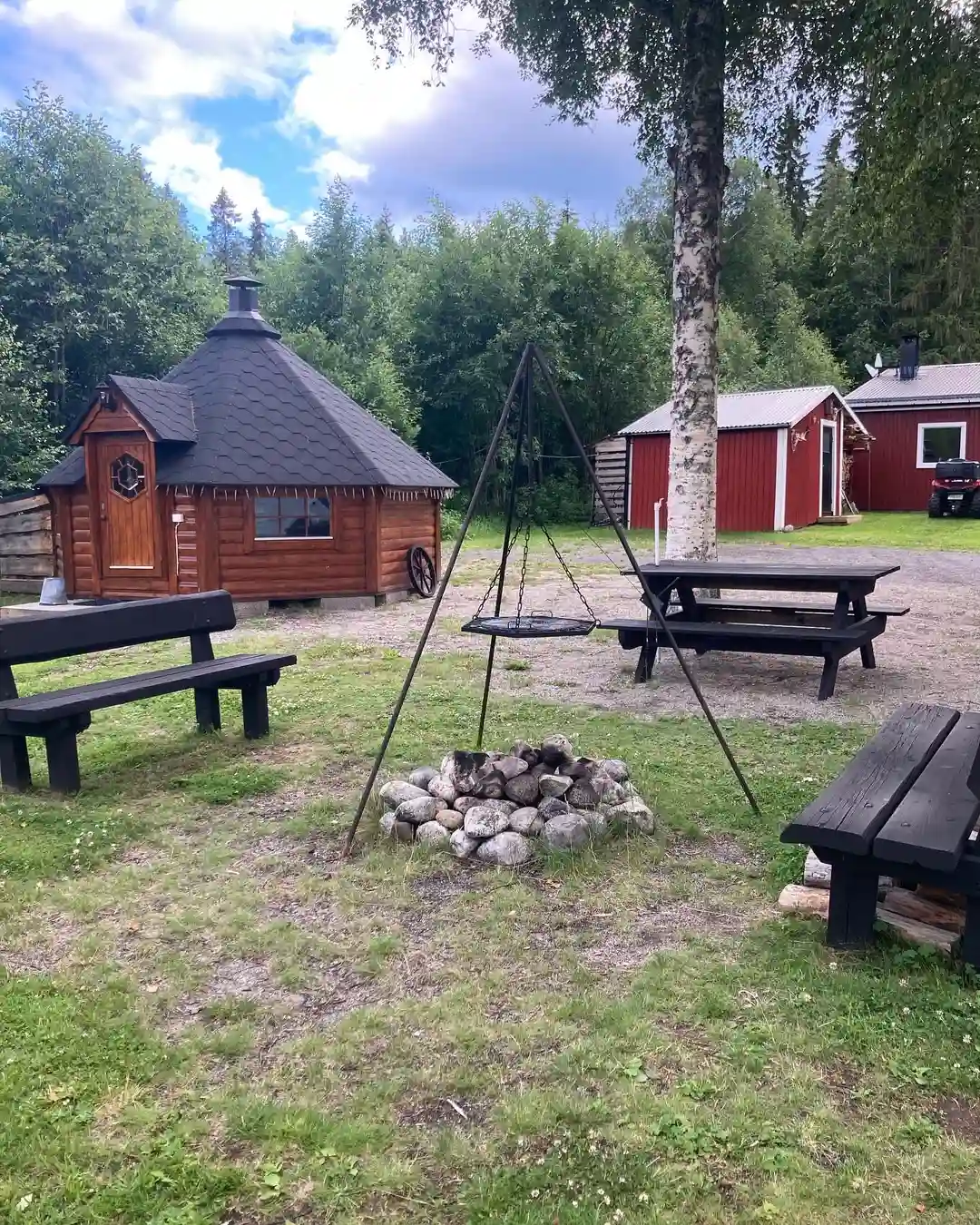 En bild av en uteplats vid Hammarstrands camping, med picknickbord och bänkar under ett solskydd. En tältstruktur syns i bakgrunden, omgiven av grönska och en parkliknande miljö.