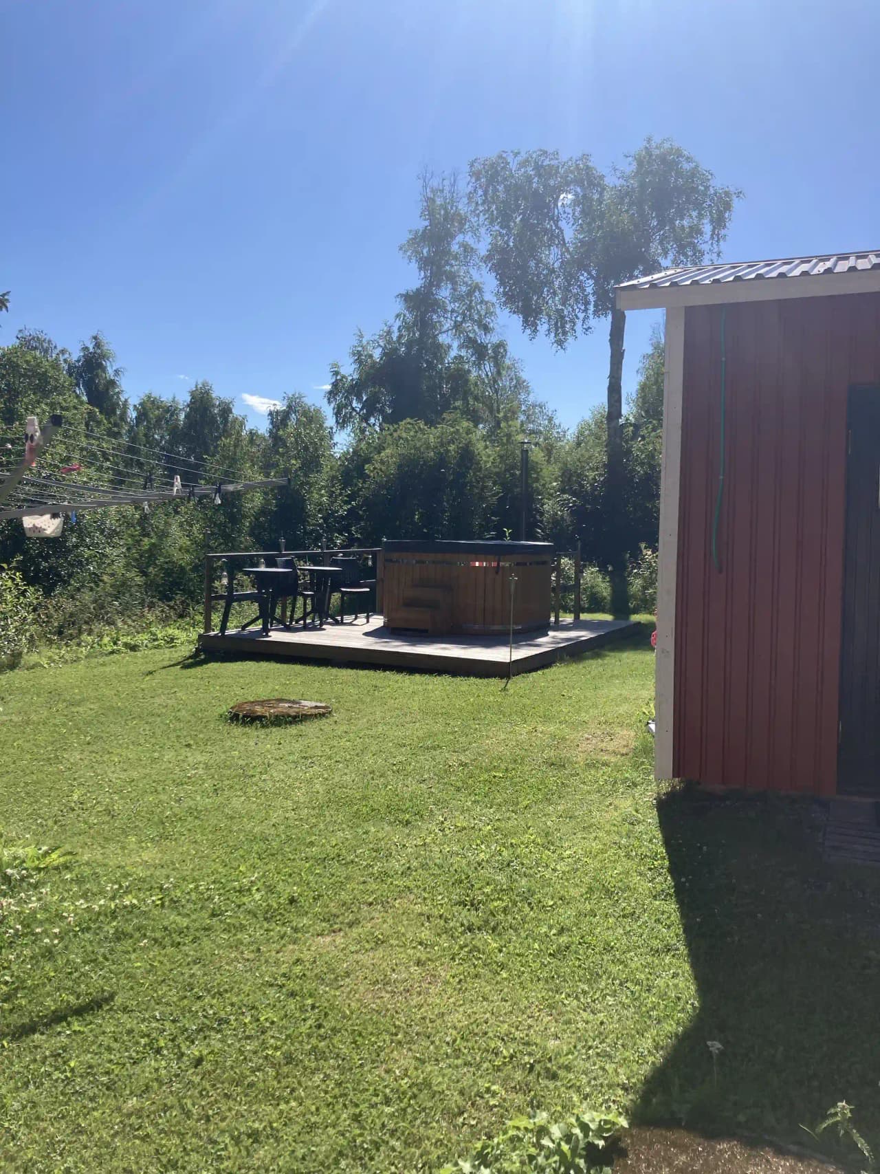 En öppen gräsyta vid Hammarstrands camping, omgiven av träd och skuggiga områden. I bakgrunden syns mindre trädgårdsbyggnader och parkmiljö.