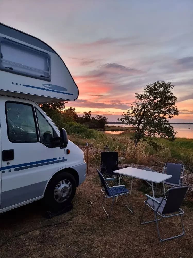 Bild av Böda Hamns Camping med husbilar och husvagnar uppställda på en campingplats. Flera campingfordon står parkerade, och i förgrunden syns hopfällbara stolar och utomhusmöbler. Solen skiner och ger en avslappnad stämning för campingbesökare.