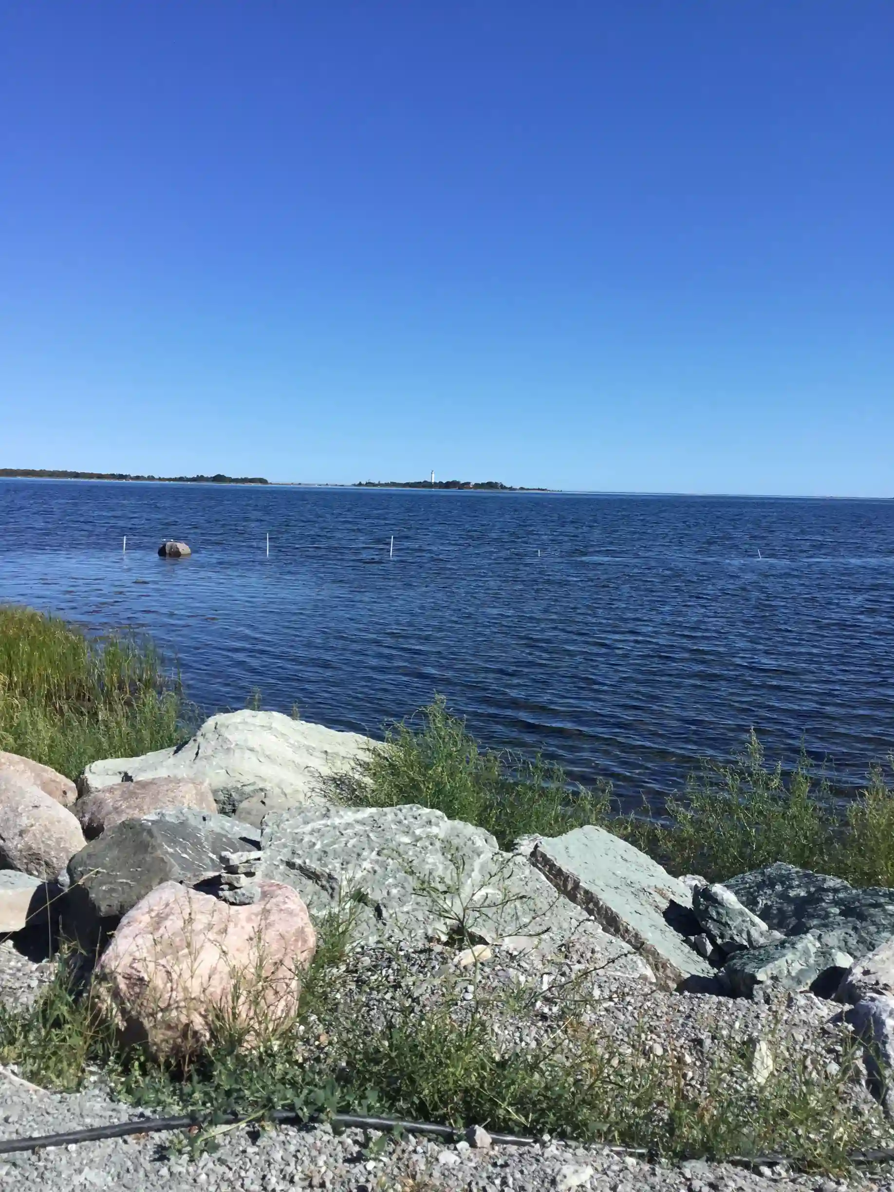 Utsikt över kustlinjen vid Böda Hamns Camping med klarblått hav, stenar längs stranden och en horisont som möter den ljusa himlen.