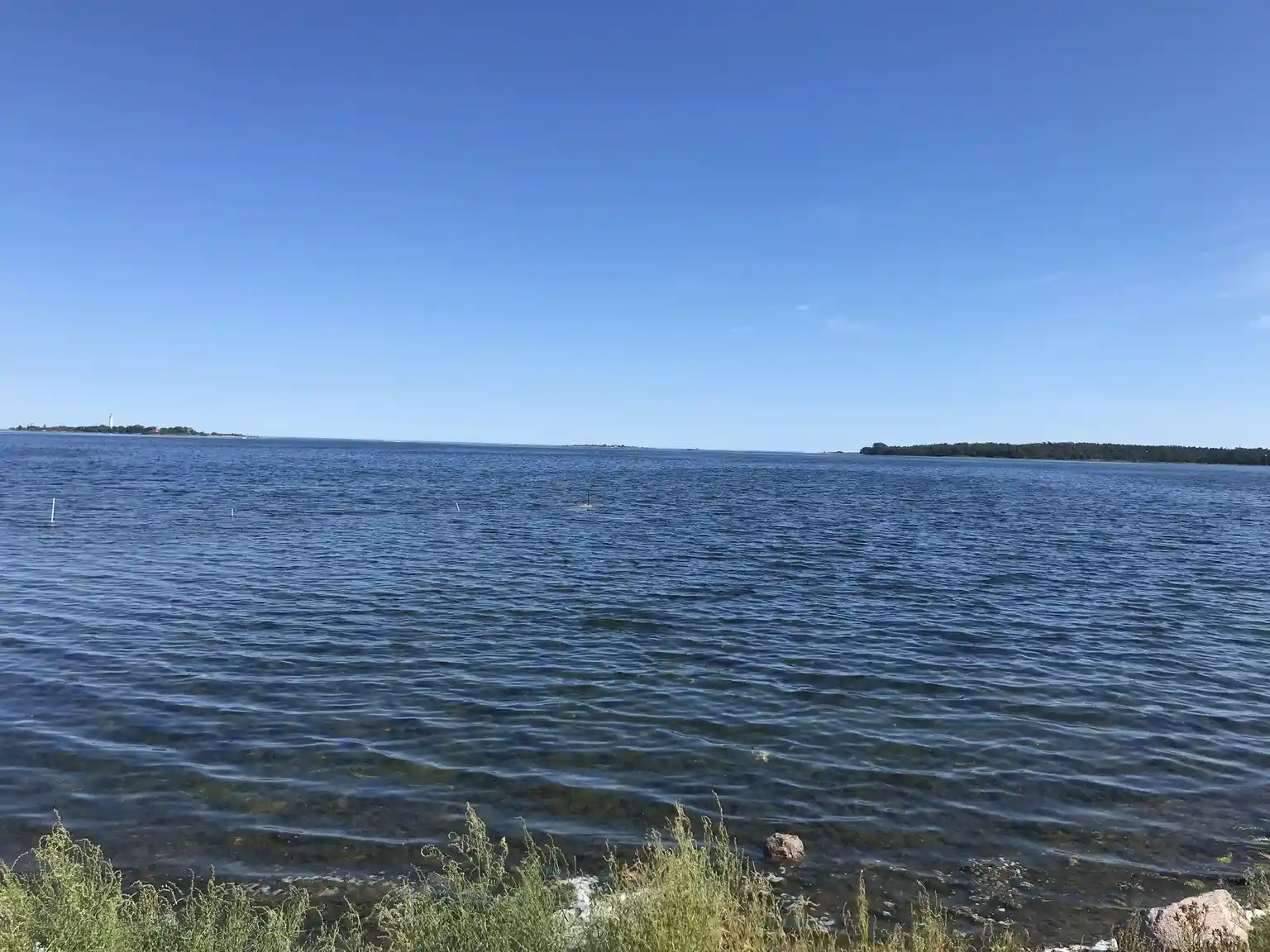 En vy över Böda hamns camping med en klarblå himmel och glittrande vatten i förgrunden, omgivande naturlandskap vid kusten.