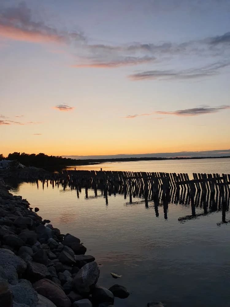 En lugn kvällsbild vid Böda Hamns Camping som visar en solnedgång över horisonten, med mjuka moln och ett spegelblankt hav som sträcker sig mot stranden.