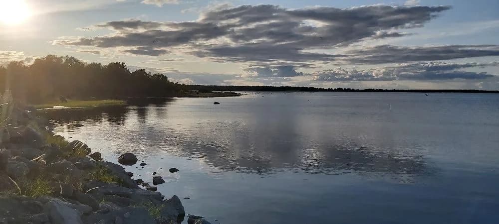 En klarblå himmel speglar sig i det stilla vattnet vid Böda Hamns Camping, där horisonten smälter samman med naturen under molnens lätt svävande formationer.