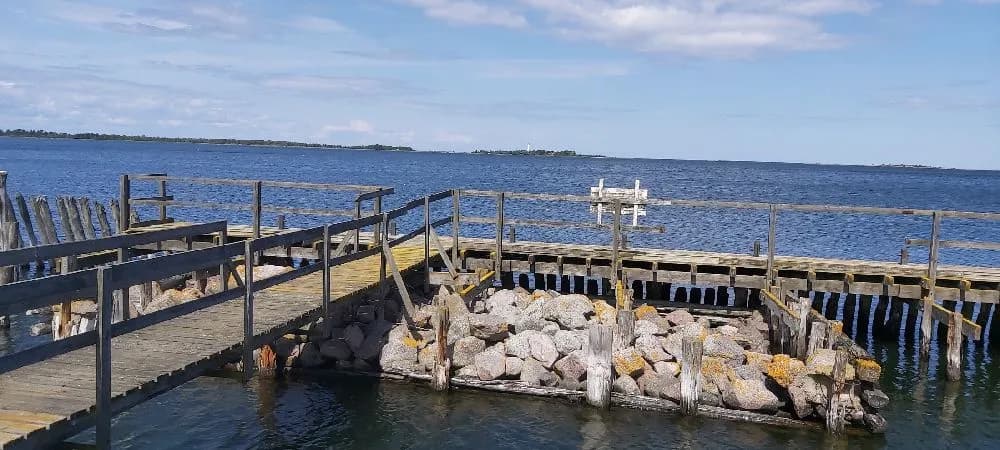 En vy över den pittoreska Böda hamns camping med en brygga som sträcker sig ut i det glittrande vattnet. I bakgrunden syns den öppna sjön omgiven av kustnära landskap.