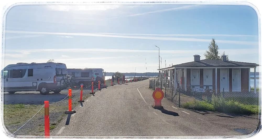 Bild på en parkeringsplats vid Böda Hamns Camping, fylld med olika fordon inklusive husbilar, skåpbilar och SUV:ar. I bakgrunden syns campingen och skogsomgivningar.