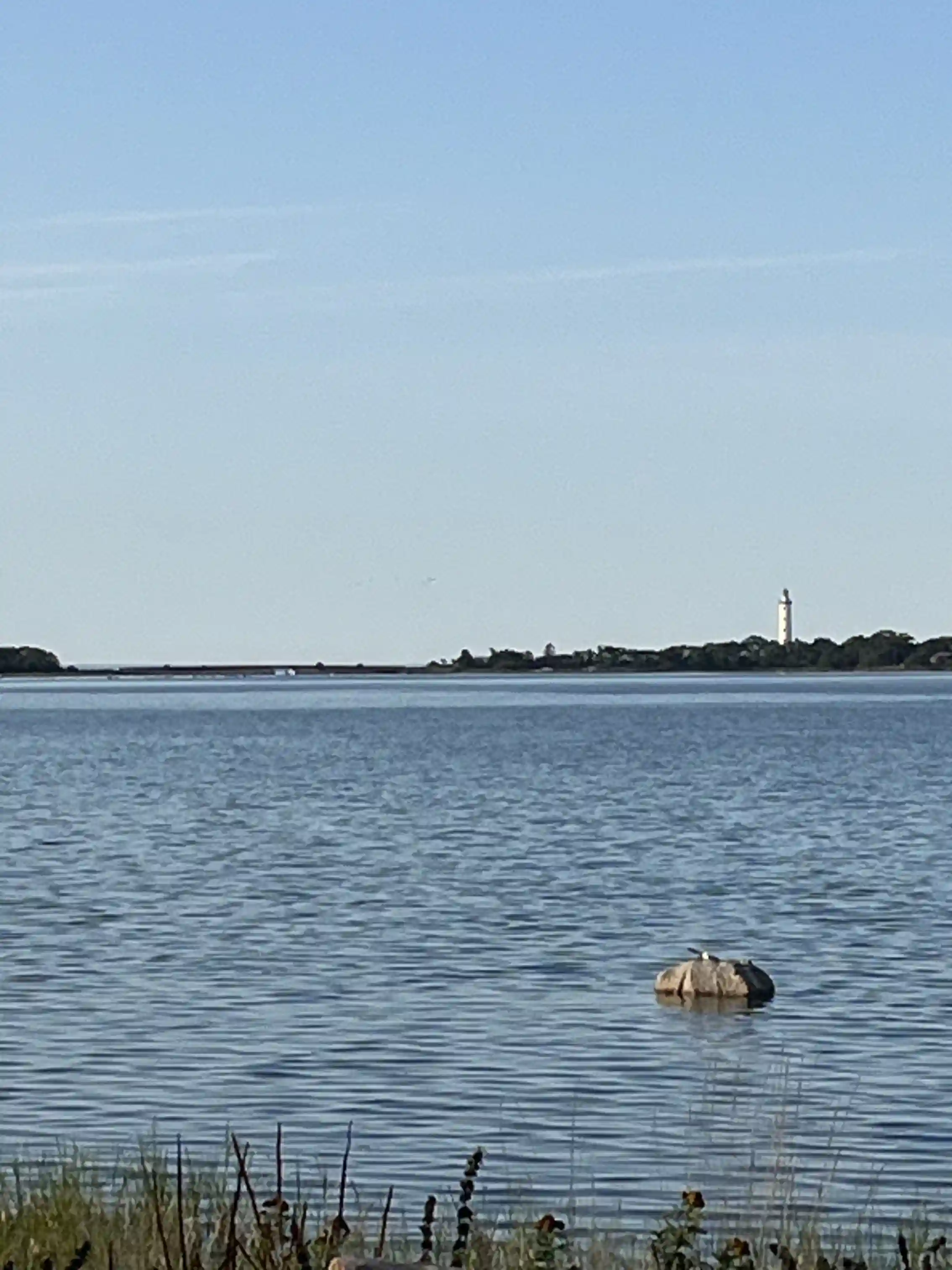 En vy över Böda hamns camping med horisonten i bakgrunden, där vatten från sjö eller hav breder ut sig. En del av en vattenväg och kustnära landskap syns, samt en byggnad eller torn på avstånd.