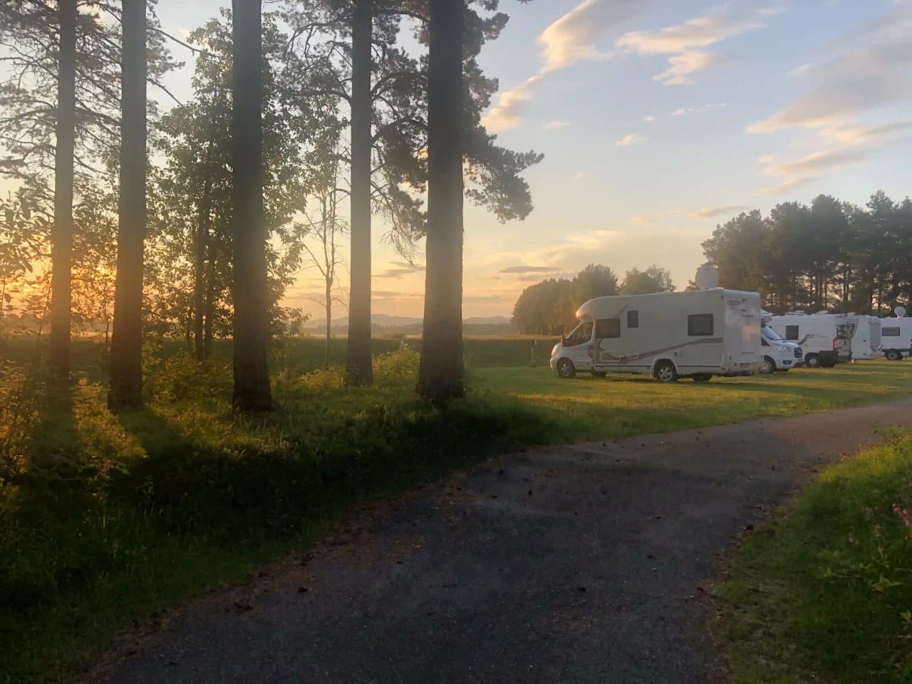 En husvagn står parkerad bland träden på Storuman camping, med soluppgången som bakgrund.