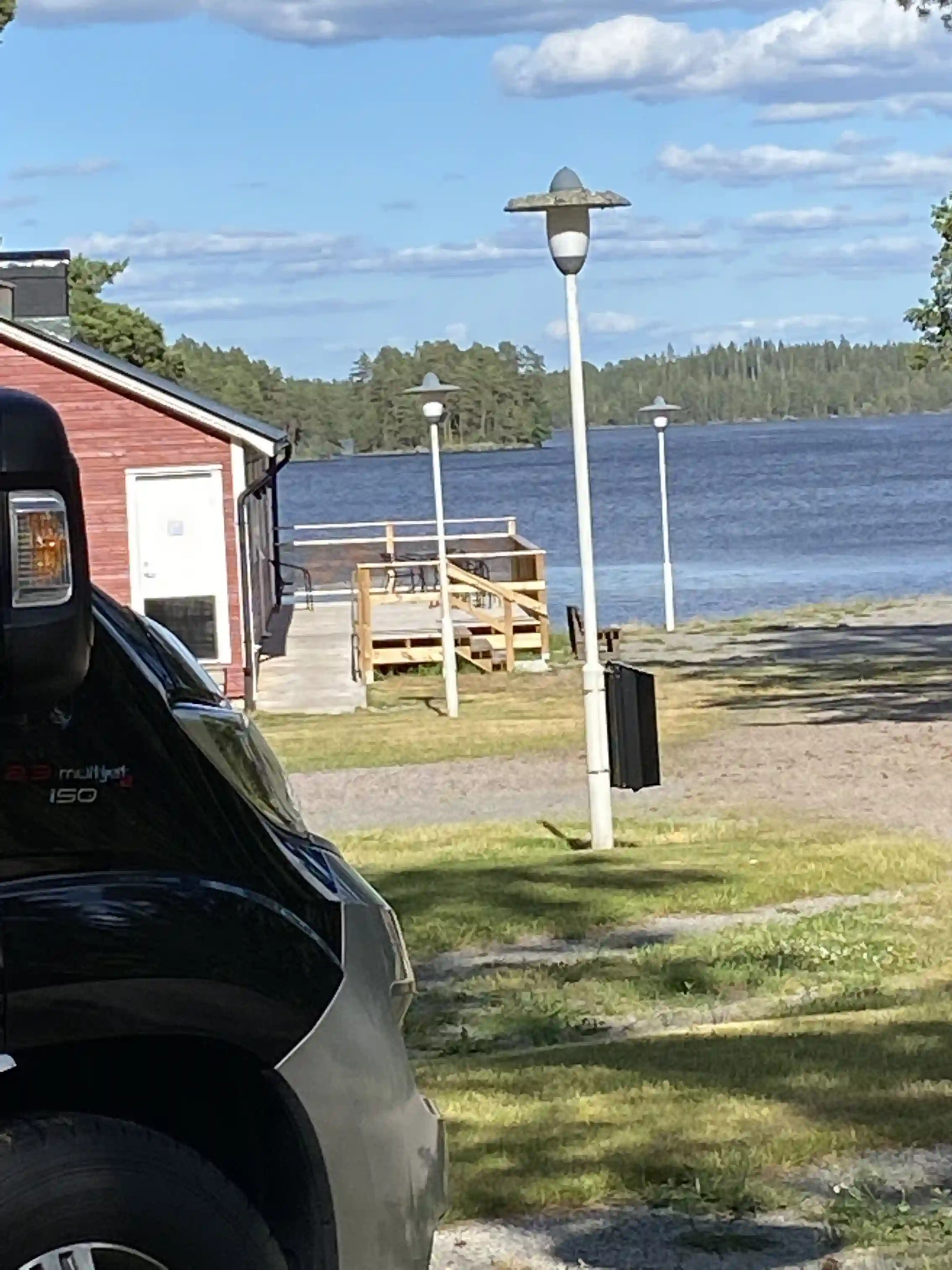 En lyxig bil står parkerad vid Uvaparken ställplats under skymningen, med en sjö och natursköna omgivningar i bakgrunden.