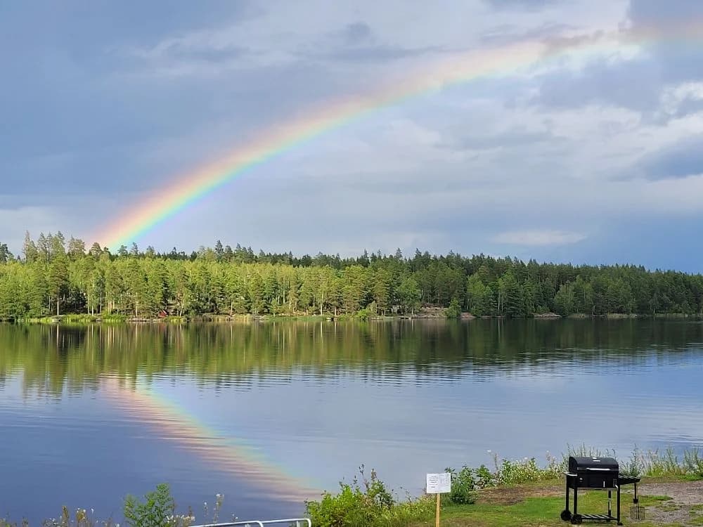 Regnbåge över en naturskön park med moln och en lugn sjö eller flod i förgrunden.
