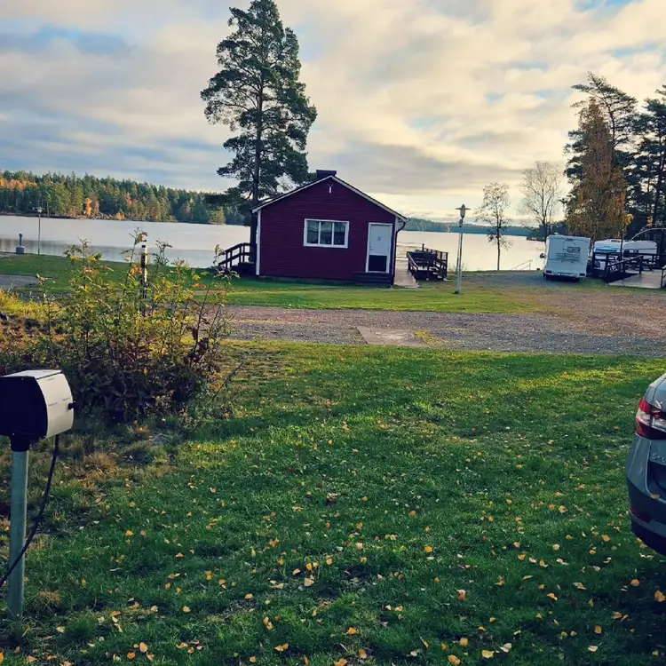 Bild av Uvaparken ställplats med en pittoresk stuga omgiven av grön gräsmatta nära en sjö. I bakgrunden syns ett naturlandskap med molnig himmel.