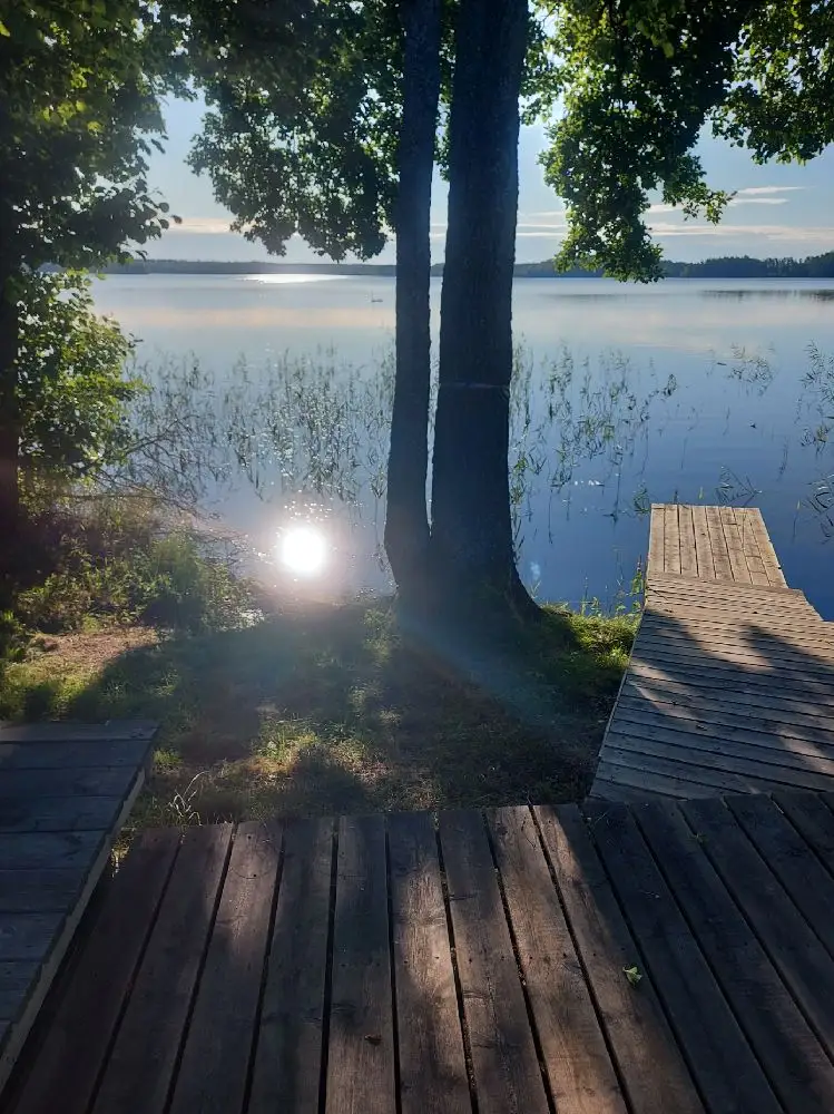 En lugn sjö vid solnedgången, med en brygga som sträcker sig ut från stranden. Vattenytan reflekterar kvällens varma ljus, omgiven av natur och grönska.