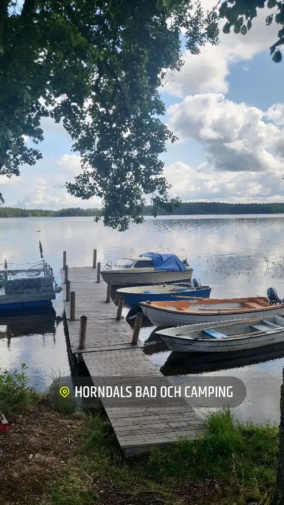 En lugn sjö med en brygga i förgrunden, där flera båtar är förtöjda. Vattnet omges av lummiga kuster under en molnig himmel. Utomhusmöbler syns längs strandkanten.