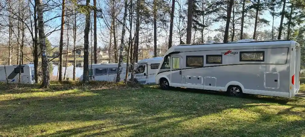En husbil och en husvagn parkerade på en naturskön campingplats i Joelskogens camping & ställplats, omgivna av träd.